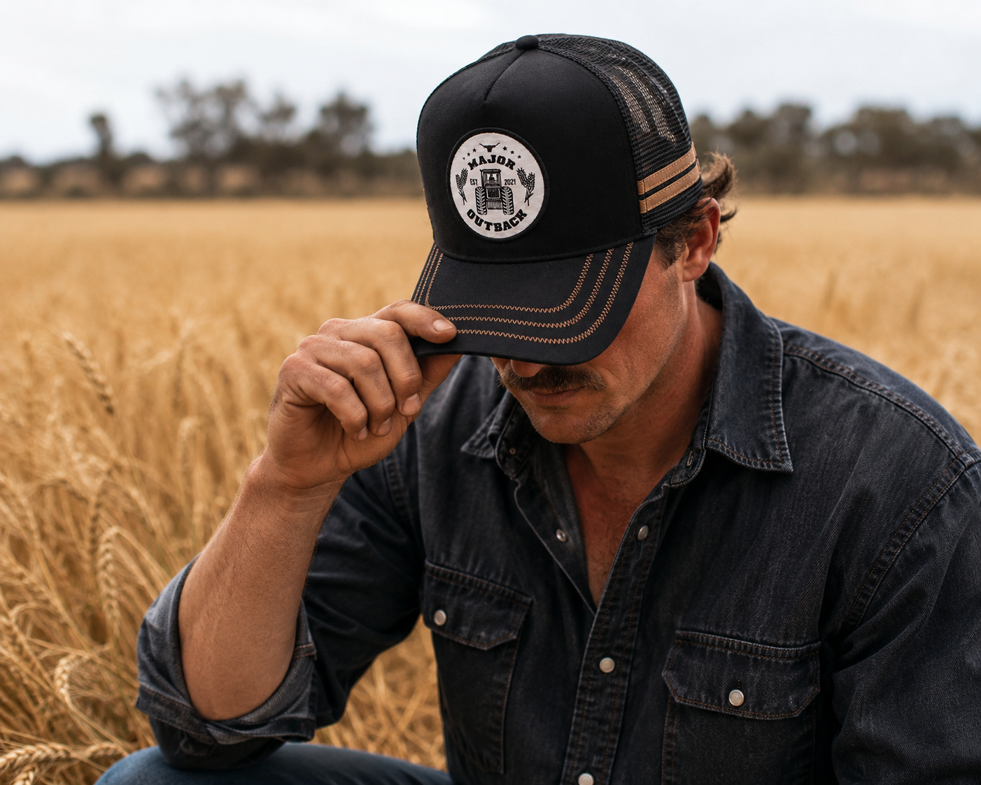 A man in a wheat field wearing a black trucker hat with a circular white and black 'Majestic Outfitter' logo patch on the front, paired with a dark denim shirt.