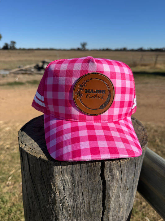 A pink and white gingham trucker hat with a brown leather patch reading 'MAJOR Outback' sits on a weathered wooden post in an outdoor field under a blue sky.