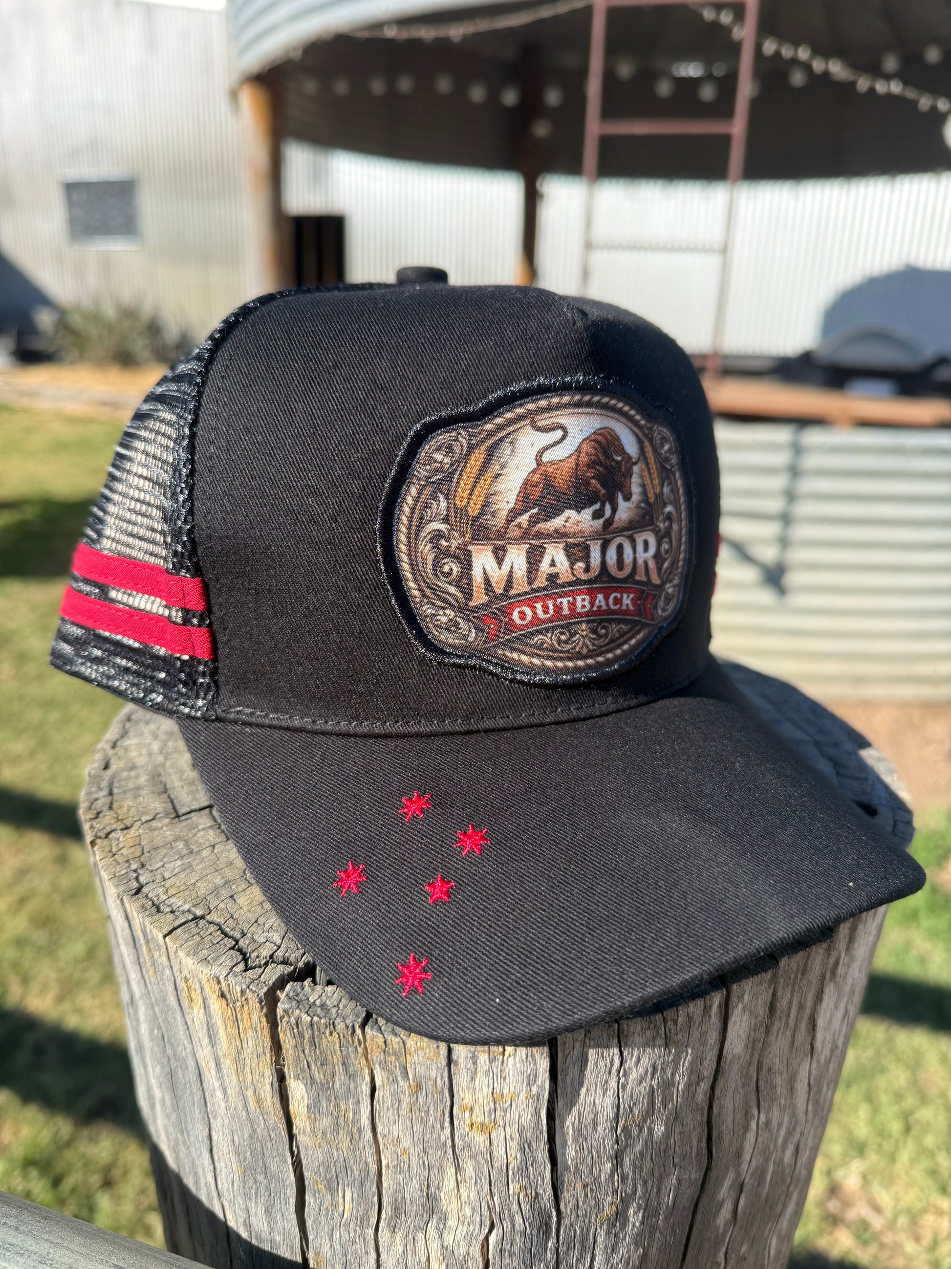 A black Major Outback trucker hat with a red and black striped side stripe and a large embroidered front patch, displayed on a weathered wooden post outdoors.