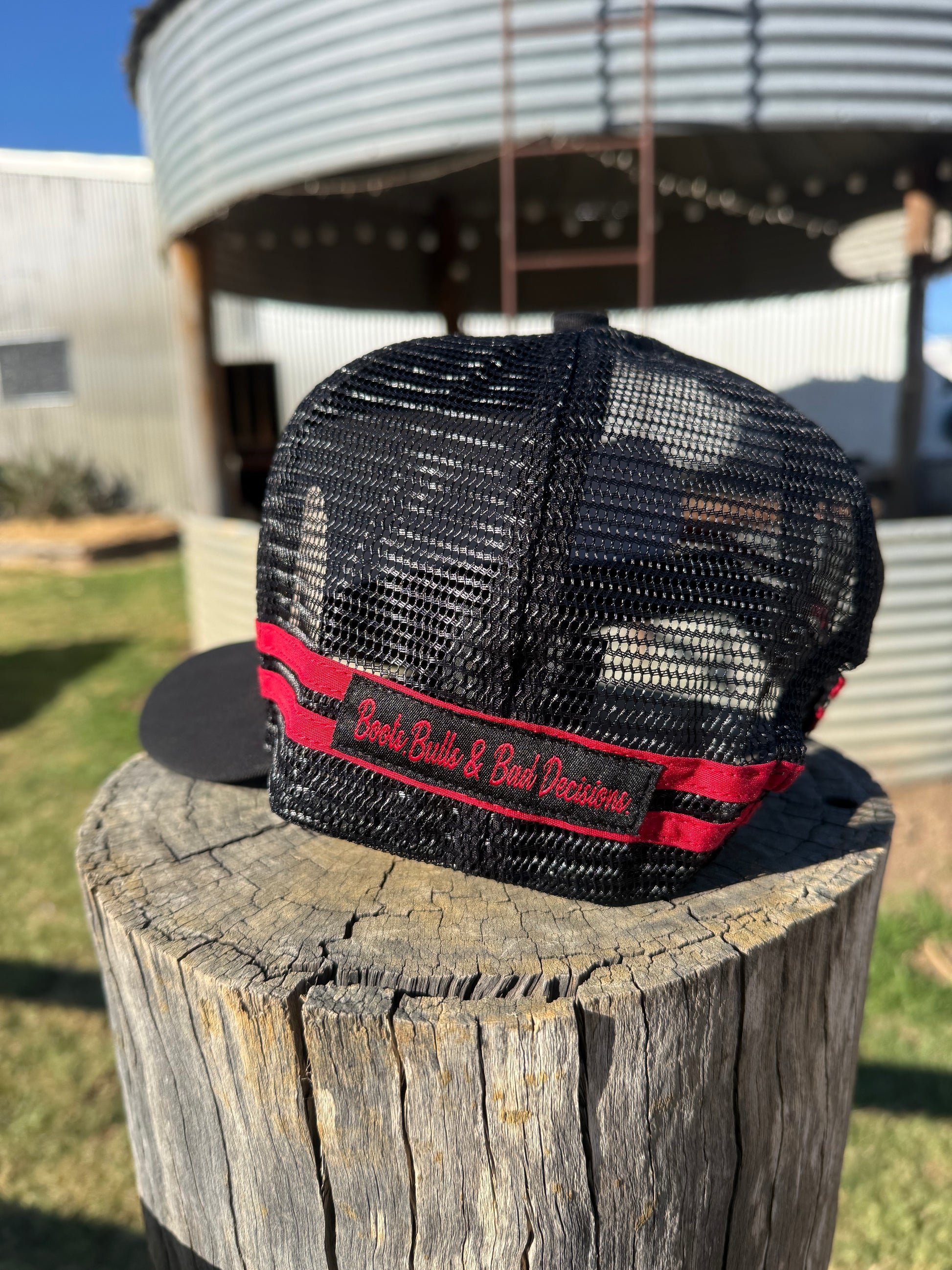 A black trucker-style cap with a red and black embroidered band reading 'Buck Bull & Buck Dealership,' displayed on a weathered wooden stump outdoors with a metal silo in the background.