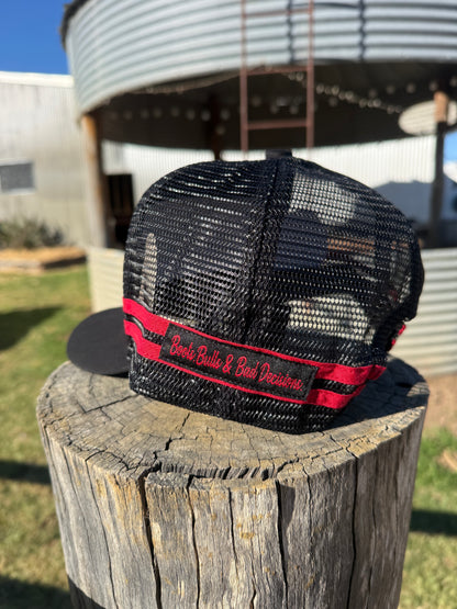 A black trucker-style cap with a red and black embroidered band reading 'Buck Bull & Buck Dealership,' displayed on a weathered wooden stump outdoors with a metal silo in the background.