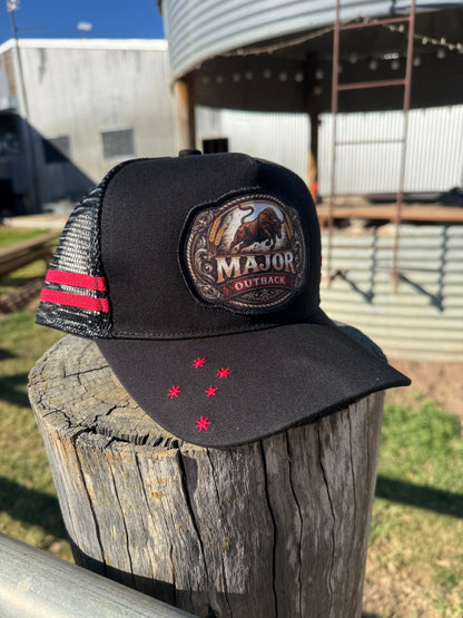 A black trucker hat with a front patch reading 'MAJOR OUTBACK' and red star accents on the brim, displayed on a weathered wooden post outdoors with a metal silo in the background.